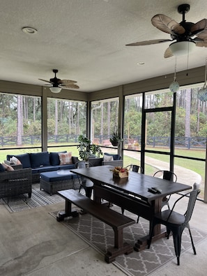Enjoy a serene view of the wooded area from the large windows in this room, which features a dining table, comfortable seating area, and a ceiling fan.