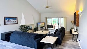 A modern living room with a sofa, a coffee table, and a TV. There is a ceiling fan, a painting on the wall, and a view of the outside through a glass door. - Sunset Rentals | Off Shore 16 (Hilton Head Island)