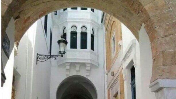 Una calle estrecha con un edificio blanco que presenta un pasaje central en arco y una torre de campanas.