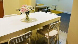 A dining area with a white table, four chairs, and a vase of flowers. A TV is mounted on the wall in the background.
