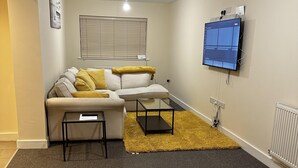 A living room with a sofa, a coffee table, a TV mounted on the wall, and a window with blinds.