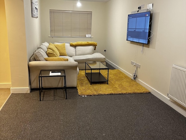 A living room with a sofa, a coffee table, a TV mounted on the wall, and a window with blinds.