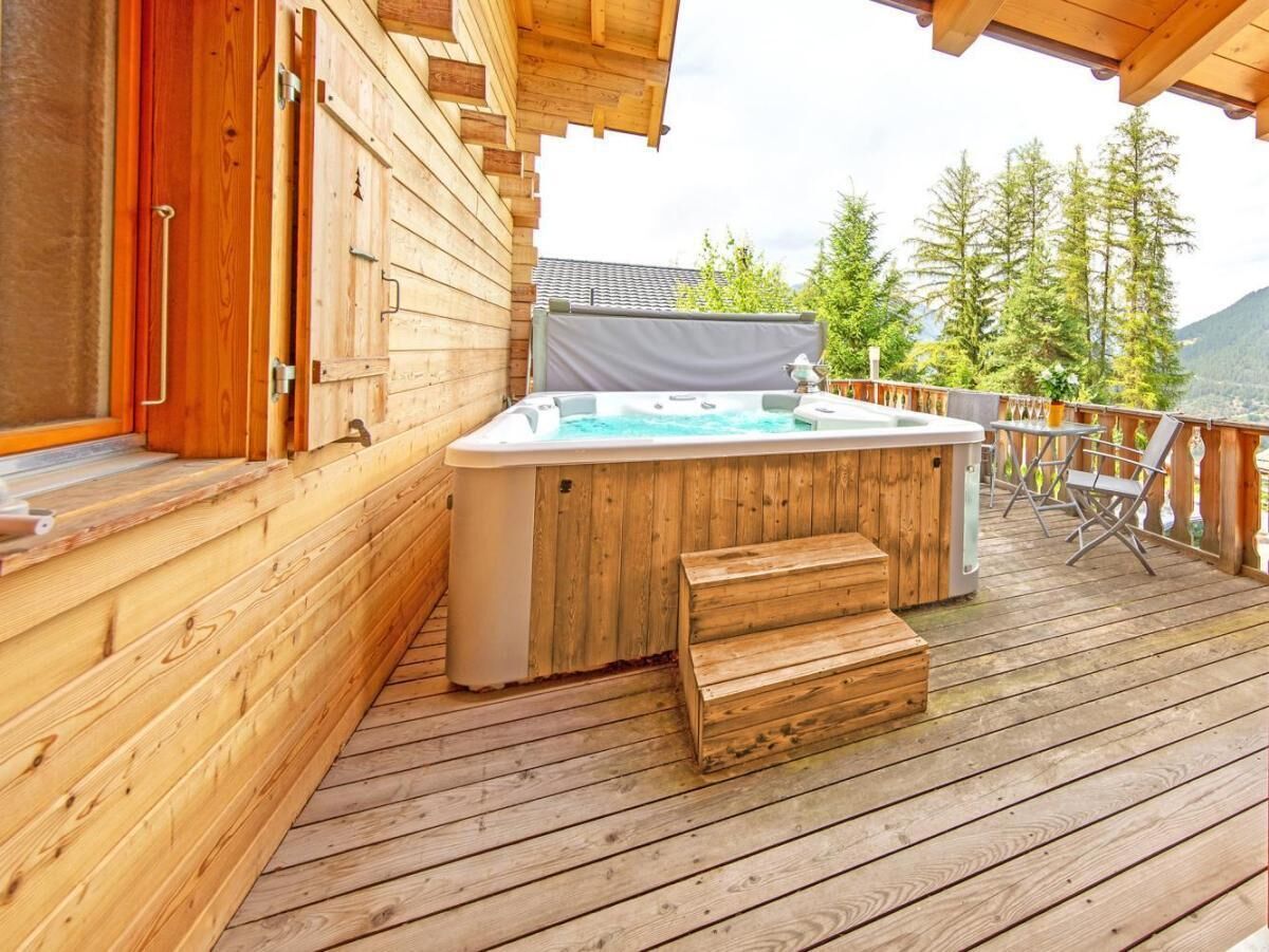 A wooden deck with a hot tub and seating area, surrounded by trees and a mountain backdrop.