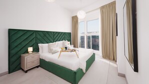 Een moderne slaapkamer met een groen bekleden hoofdeinde, een nachtkastje met een lamp en een groot raam met gordijnen.
