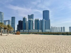 Une plage de sable bordée de palmiers, avec la silhouette d’une ville en arrière-plan.