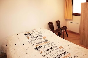 A bedroom with a bed covered in a polka dot duvet, two wooden chairs, a window with yellow curtains, and a radiator.