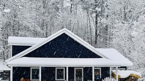 A two-story house with a dark exterior and white trim, surrounded by snow-covered trees. - Charming Cavendish Guesthouse (Cavendish)