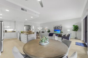 A modern kitchen with white cabinets, a dining area with a round table, and a living room with a TV and seating.