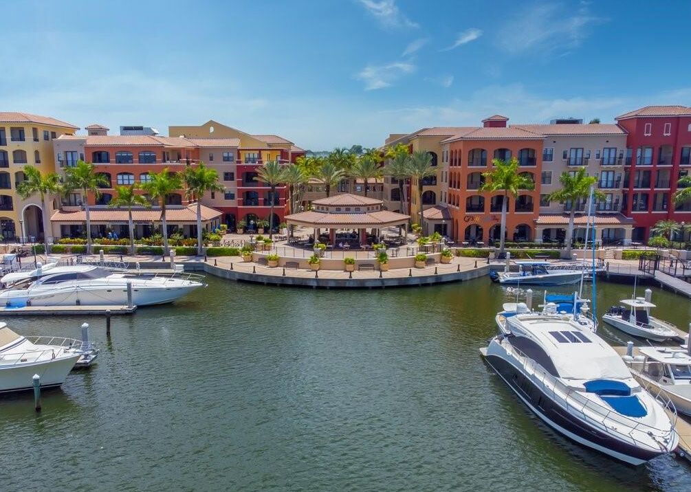 A marina with boats docked, surrounded by multi-story buildings with balconies and palm trees.