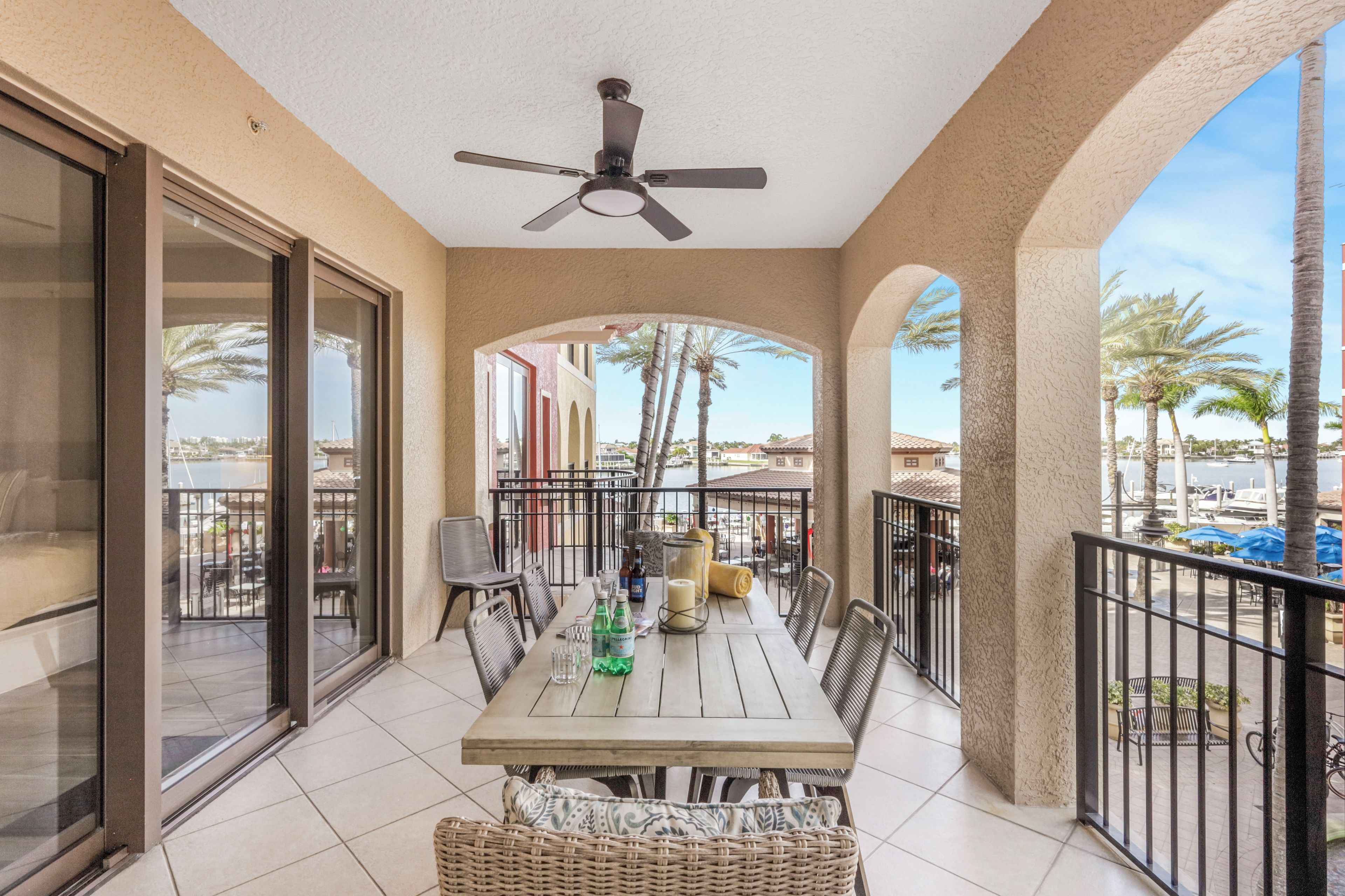 A balcony with a table set for dining, a ceiling fan, and a view of palm trees and buildings.