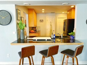 Una cocina moderna con una isla central, dos taburetes de bar, armarios de madera, un refrigerador y un reloj en la pared.