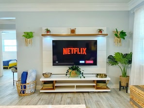 Una sala de estar moderna con un televisor montado que muestra el logo de Netflix, un mueble blanco con estantes y varias plantas en macetas.