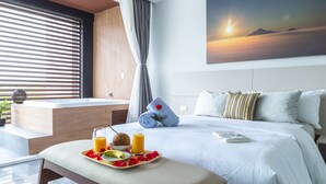 A hotel room with a bed, a breakfast tray with fruit and juice, a painting of a sunset, and a view of the outdoors through the window.