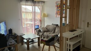 Una sala de estar con un sofá, una mesa de centro, un televisor y una computadora portátil sobre un escritorio. Hay una ventana con cortinas, una lámpara y un cuadro en la pared.