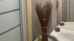Un baño con lavamanos blanco, un espejo y un jarrón con plantas secas sobre una estantería pequeña.