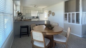 Un coin repas avec une table ronde en bois et quatre chaises, une cuisine en arrière-plan et une vue à travers une fenêtre avec des stores.