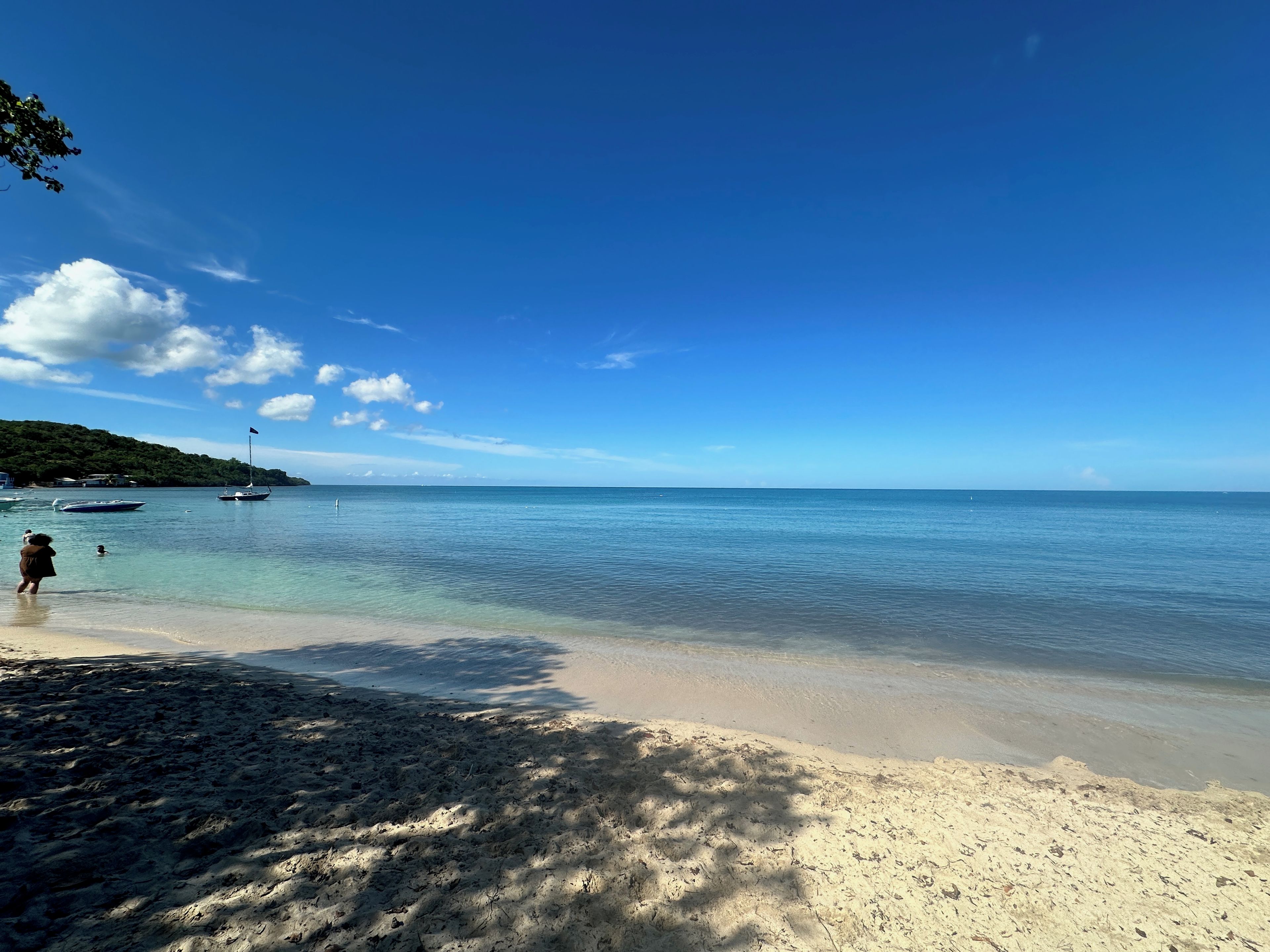 A beach with clear water, boats, and a person walking.