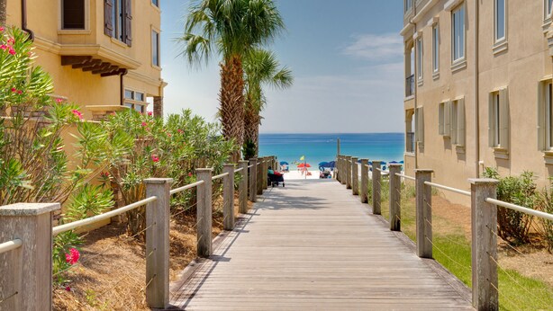 Een houten pad dat naar een strand leidt, met een palmpijlstaart en een gebouw met balkons.