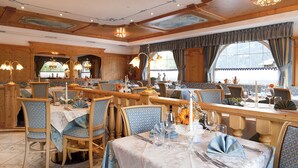 Un espace repas comprenant plusieurs tables et chaises, des poutres en bois au plafond et de grandes fenêtres avec des rideaux.