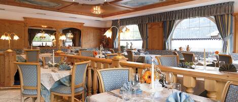 A dining area with multiple tables and chairs, wooden beams on the ceiling, and large windows with curtains.