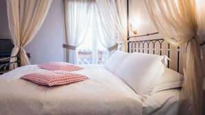 Un lit bien fait, avec du linge de lit blanc et des coussins à motifs rouges, une tête de lit en métal et des rideaux légers, attachés pour laisser apparaître un balcon.
