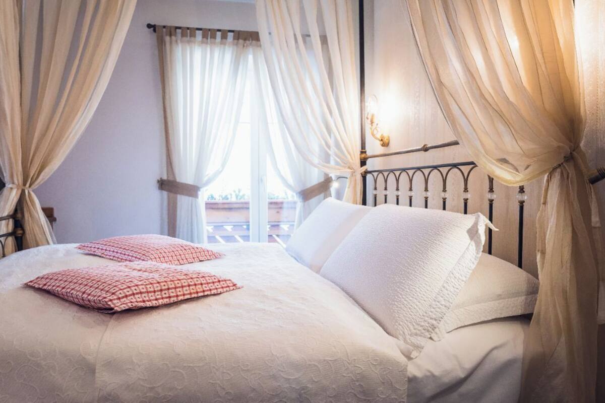 Un lit bien fait, avec du linge de lit blanc et des coussins à motifs rouges, une tête de lit en métal et des rideaux légers, attachés pour laisser apparaître un balcon.