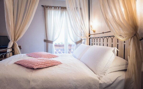 Una cama bien hecha con sábanas blancas y almohadas rojas estampadas, cabecera de metal y cortinas traslúcidas atadas para mostrar un balcón.