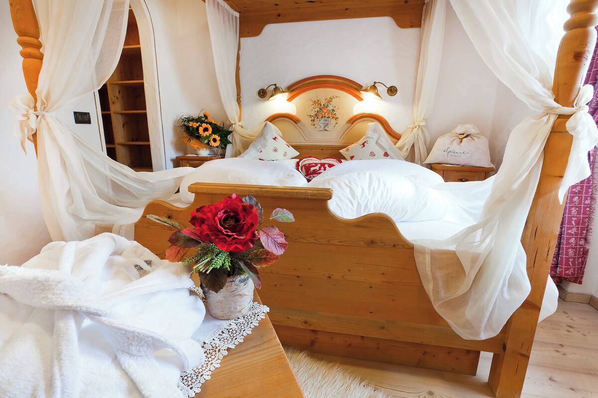 Un lit en bois avec une literie blanche et un baldaquin, un vase rempli de fleurs et une petite table recouverte d’une nappe en dentelle.