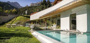 A modern building with a swimming pool, outdoor seating, and a view of mountains. - Renato Hotel (San Giovanni di Fassa)