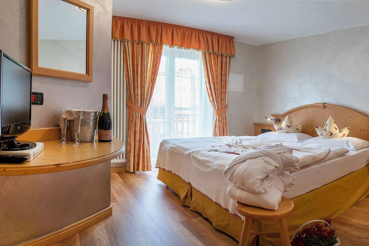 Une chambre d’hôtel avec un lit, une table de chevet en bois, une chaise, une petite table, un miroir et une fenêtre avec des rideaux.