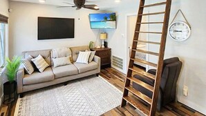 A living room with a sofa, a TV mounted on the wall, a ceiling fan, and a wooden ladder against the wall.