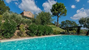 A swimming pool with clear blue water, surrounded by lush greenery and a hillside with a building. - Casa Antonio - Private villa, 4 bedrooms, pool, A/C, wifi, pets, Marche (Mogliano)