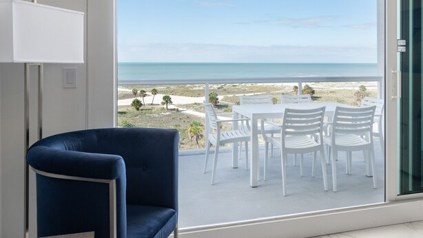 Een moderne kamer met een groot raam dat uitkijkt over een strand. Er staat een blauwe stoel en een witte tafel met stoelen.