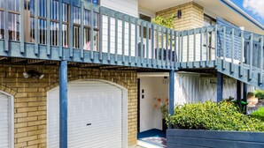 A two-story residential building with a blue balcony, a garage door, and a small garden area with potted plants. - Coastal Retreat Unit close to Beach (Auckland North Shore)