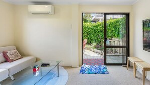 A modern living room with a glass coffee table, a sofa with a cushion, a bench, and a sliding glass door leading to an outdoor area with a patterned rug. - Coastal Retreat Unit close to Beach (Auckland North Shore)