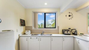 A kitchen with white cabinets, a sink, a window, and a clock on the wall. - Coastal Retreat Unit close to Beach (Auckland North Shore)