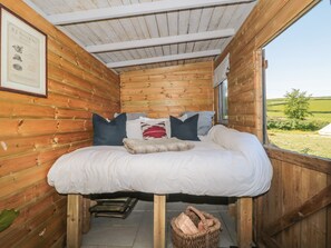 A wooden cabin bedroom with a bed, pillows, a framed certificate on the wall, and a window overlooking a green landscape. - The Snug (Dulverton)