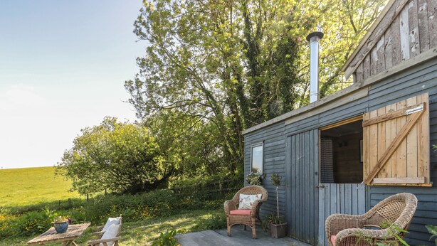A rustic wooden cabin with a porch, wicker chairs, and a small table. - The Snug (Dulverton)