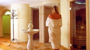 A woman in a robe standing in a bathroom with a shower, a decorative fountain, and a sign that reads "DOCCIA". - Hotel Alle Alpi (Moena)