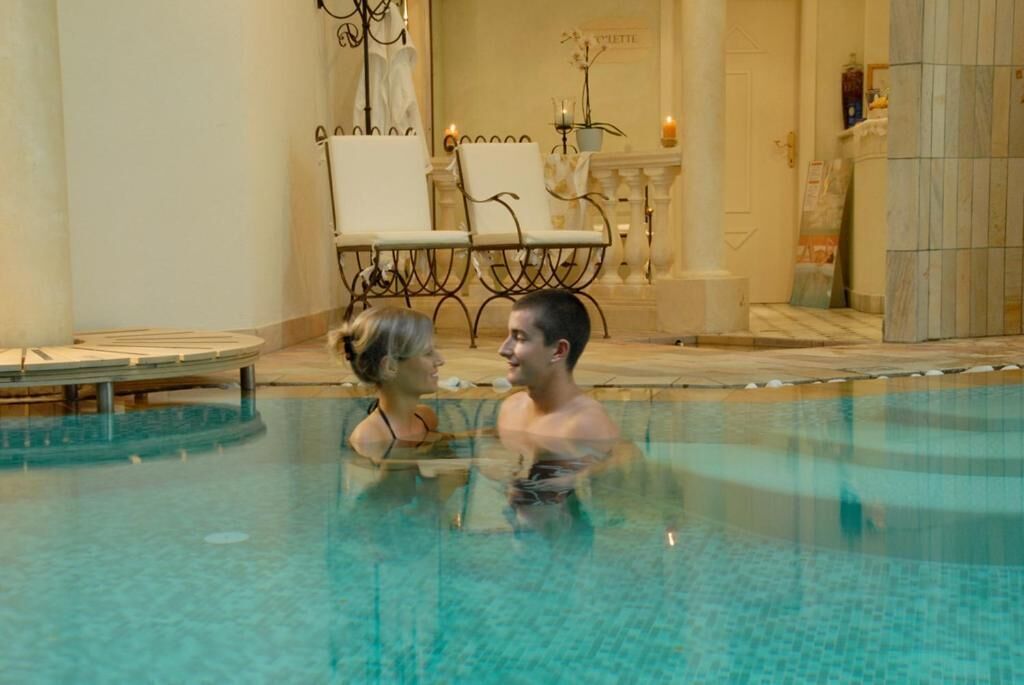 A couple in a clear indoor pool with a lounge area in the background.
