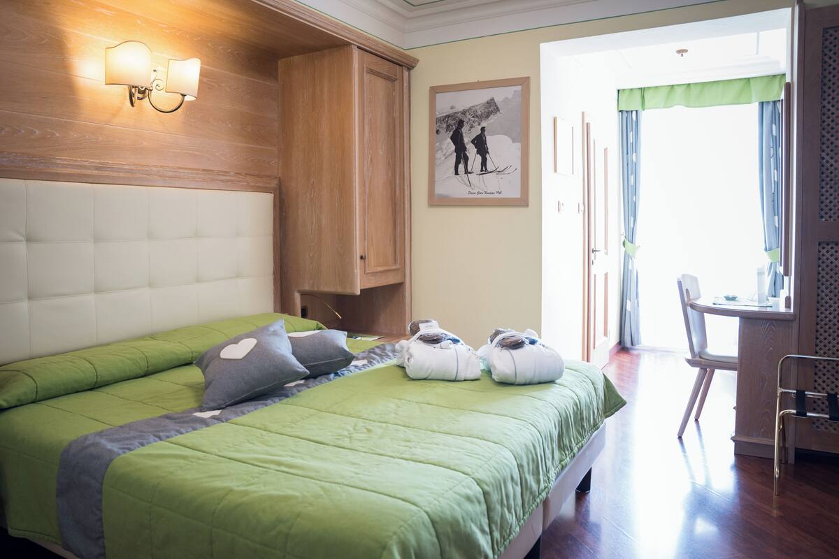 A hotel room with a bed, a wooden wardrobe, a desk with a chair, and a framed picture on the wall.