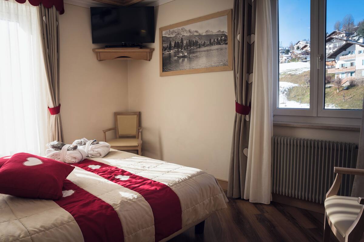 A hotel room with a bed, a chair, a TV, a window with a view of a lake and mountains, and a radiator.