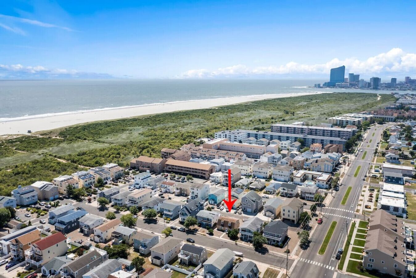 Vista aérea de una zona residencial costera con casas unifamiliares y edificios de apartamentos, una playa al fondo y una flecha roja destacada que apunta a un edificio específico.