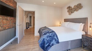 Un dormitorio moderno con pared de ladrillo, una cama grande con ropa de cama blanca, una manta azul y un televisor montado en la pared.