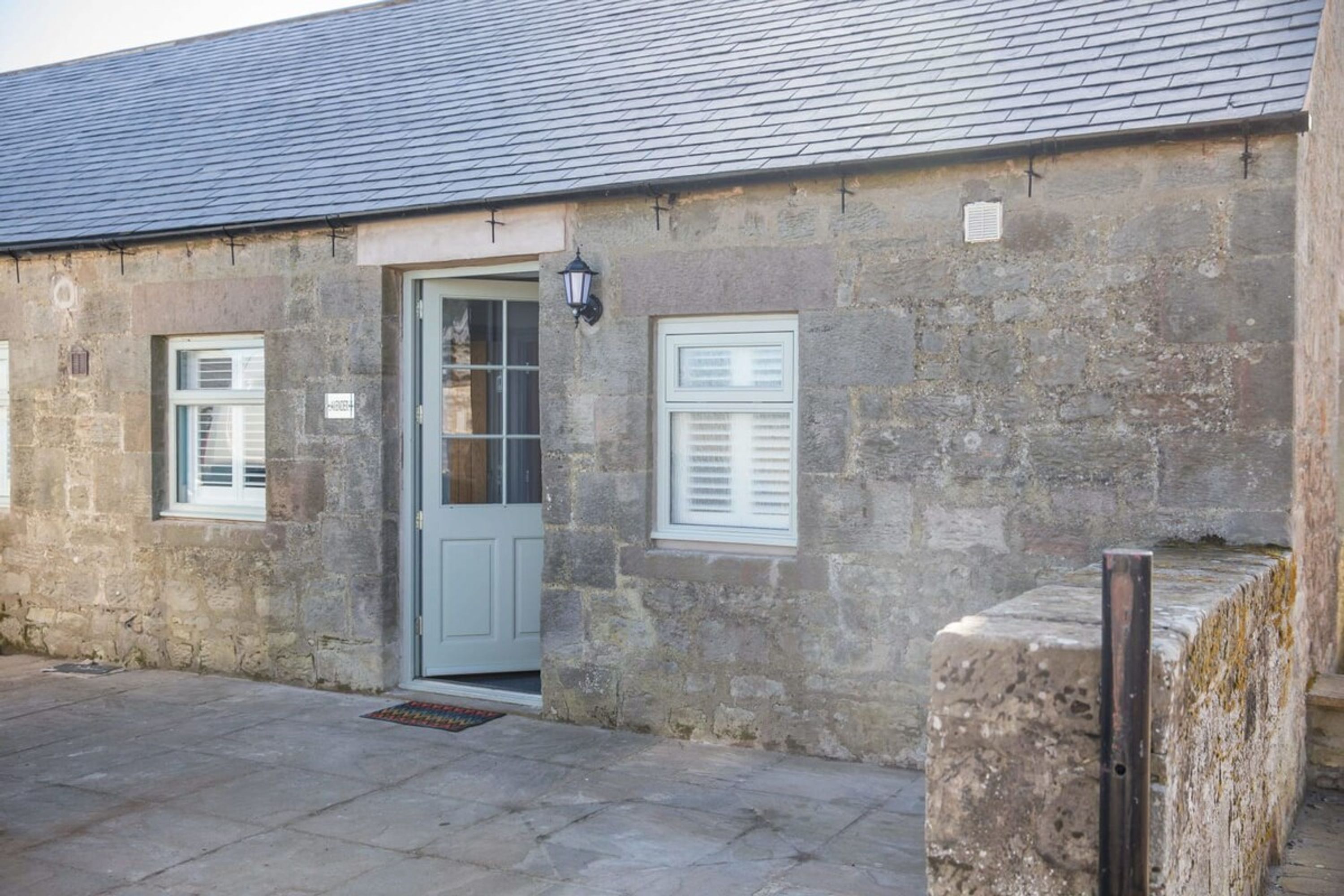 A stone building with a grey door, two windows with shutters, and a small lamp.