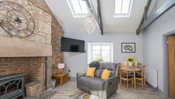 A living room with a fireplace, a sofa, a TV mounted on the wall, and a dining area with a table and chairs. - Host Stay No 2 Lavender Cottage (Belford)