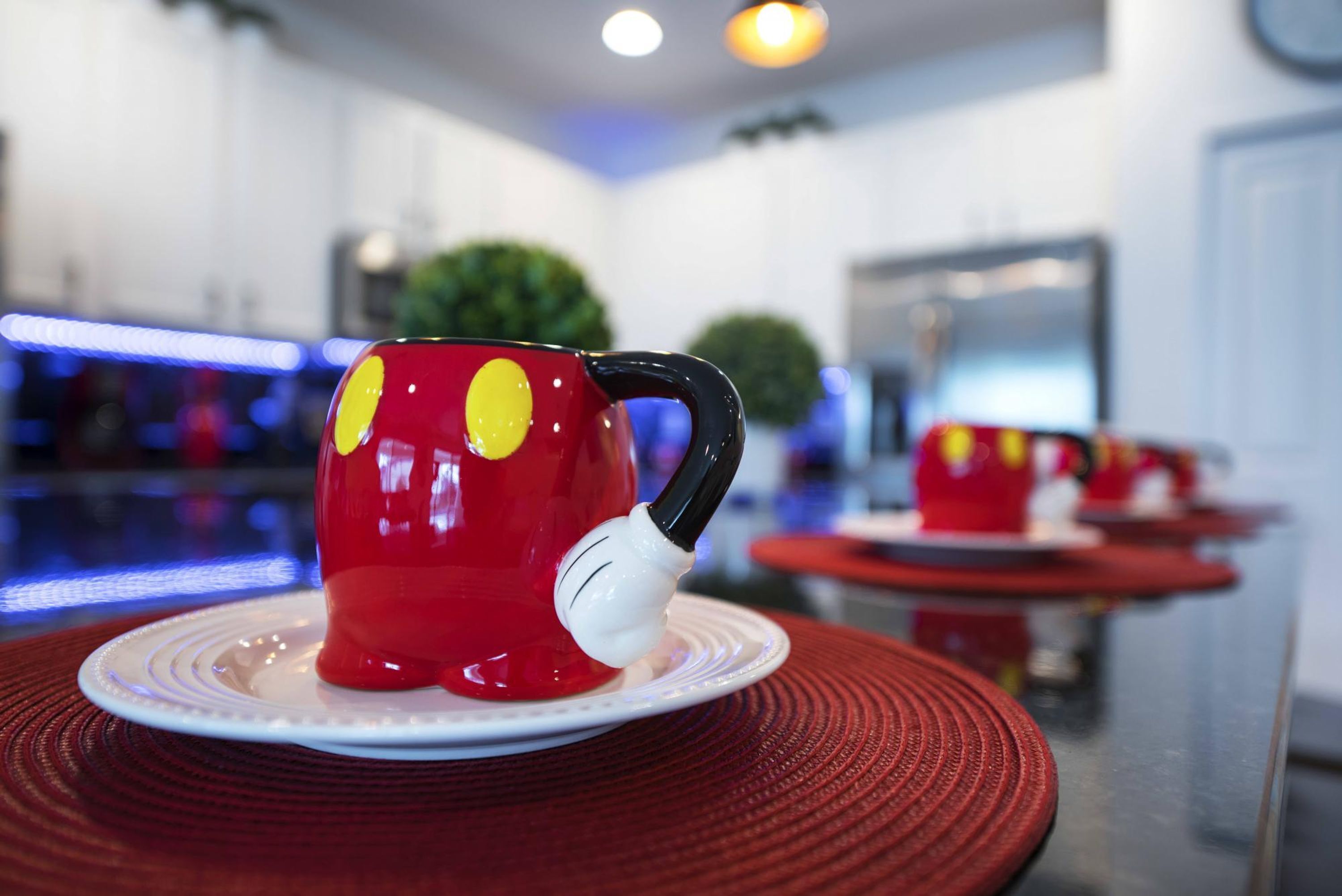 Una taza roja con asa blanca sobre un plato blanco, colocada sobre una mesa roja en una cocina moderna.