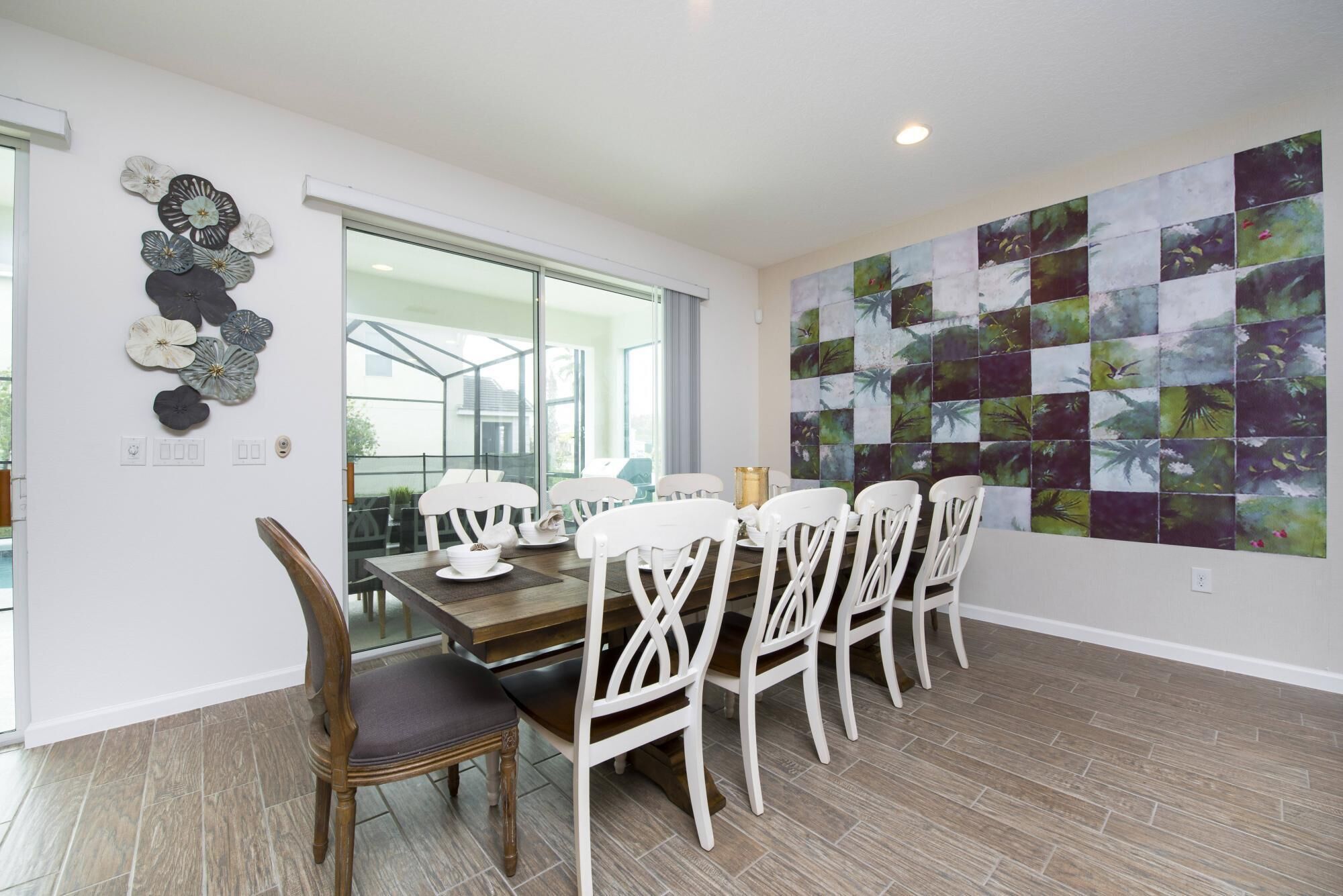 A dining area with a wooden table and white chairs, a glass door leading outside, and a wall with a floral tile mural.