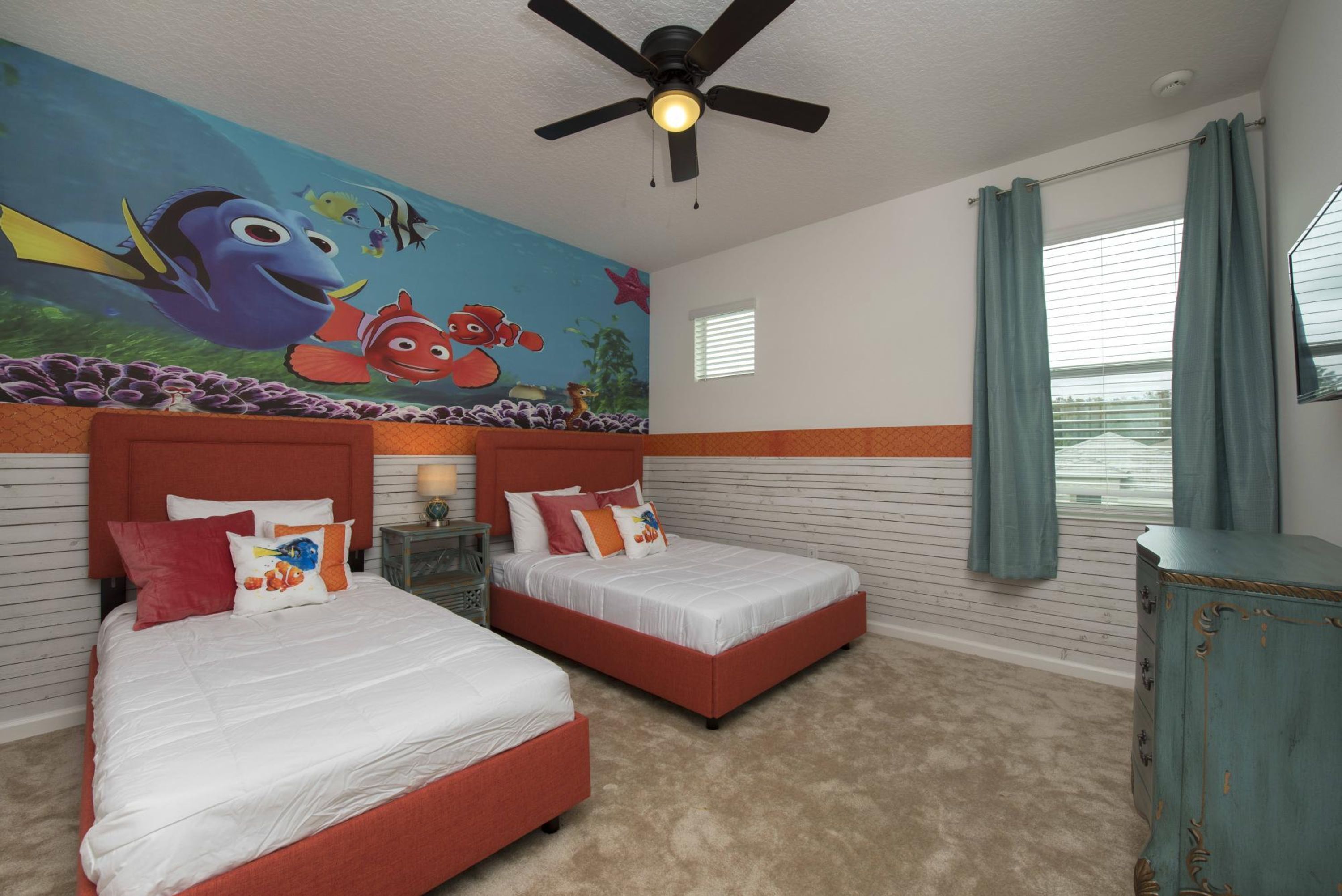 Une chambre avec deux lits, un ventilateur de plafond, une fresque représentant des poissons et une fenêtre avec des rideaux.