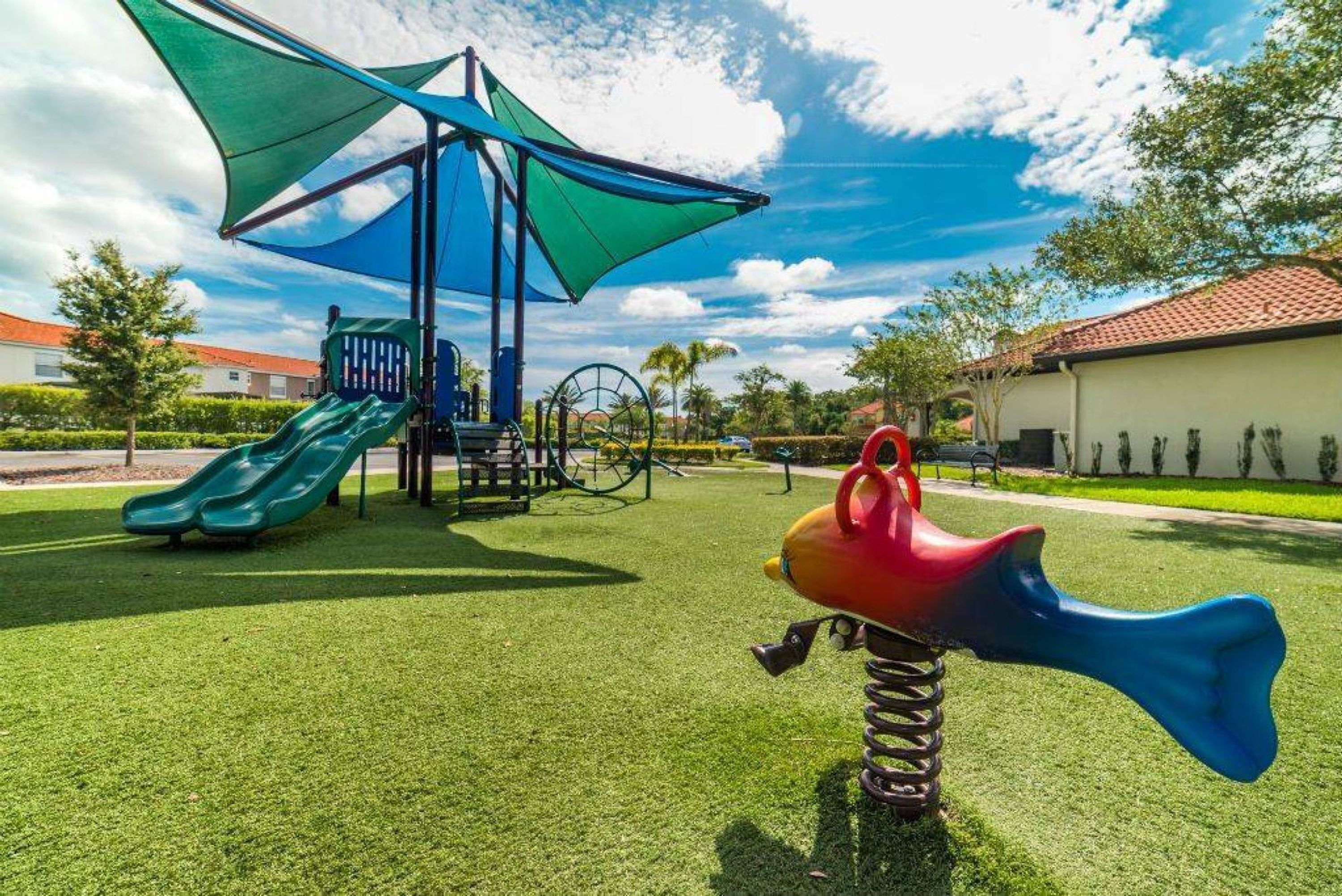 A playground with a slide and swings, surrounded by green grass and a blue canopy.